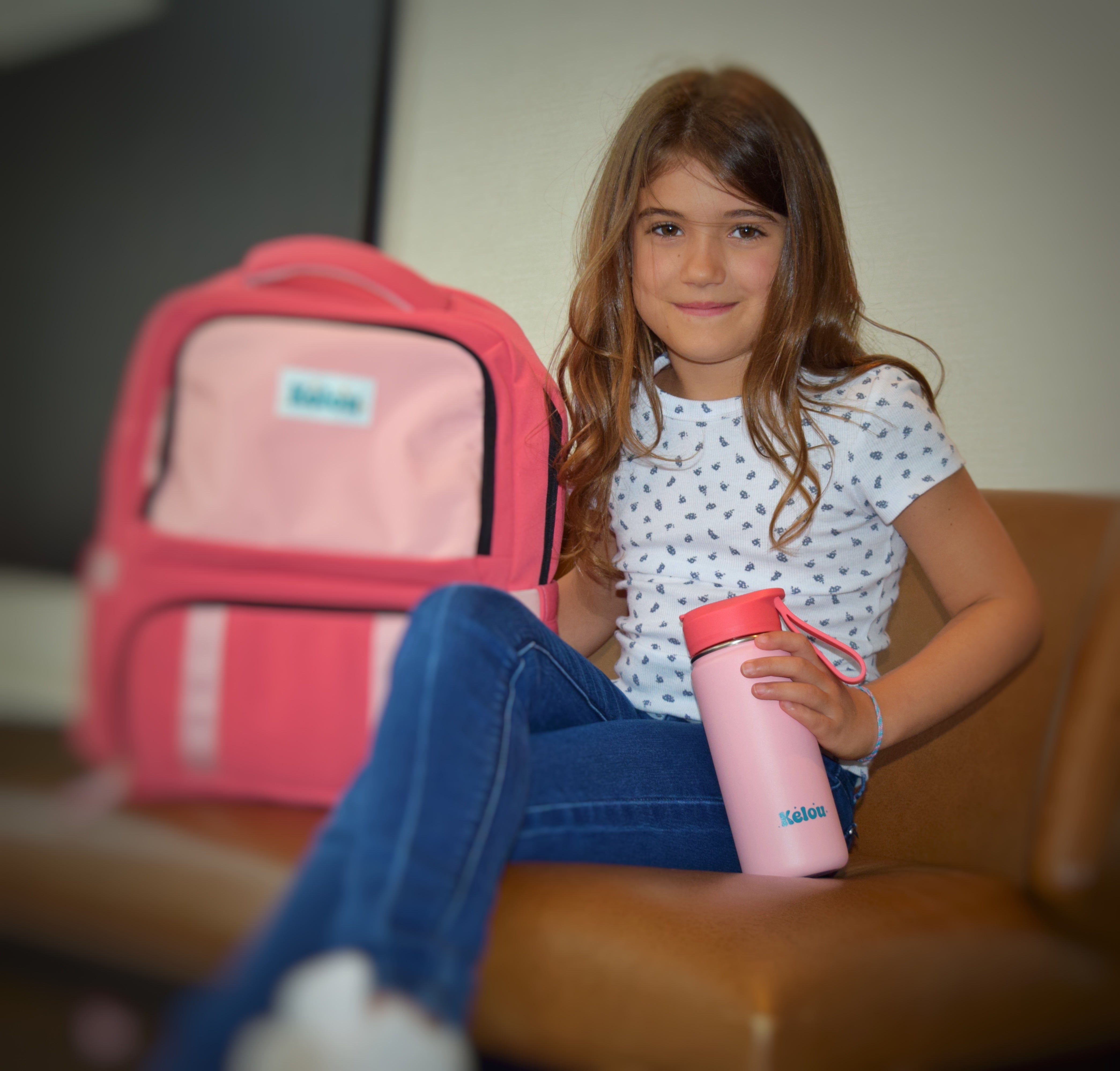 Young girl sitting on a couch with a pink backpack and water bottle.