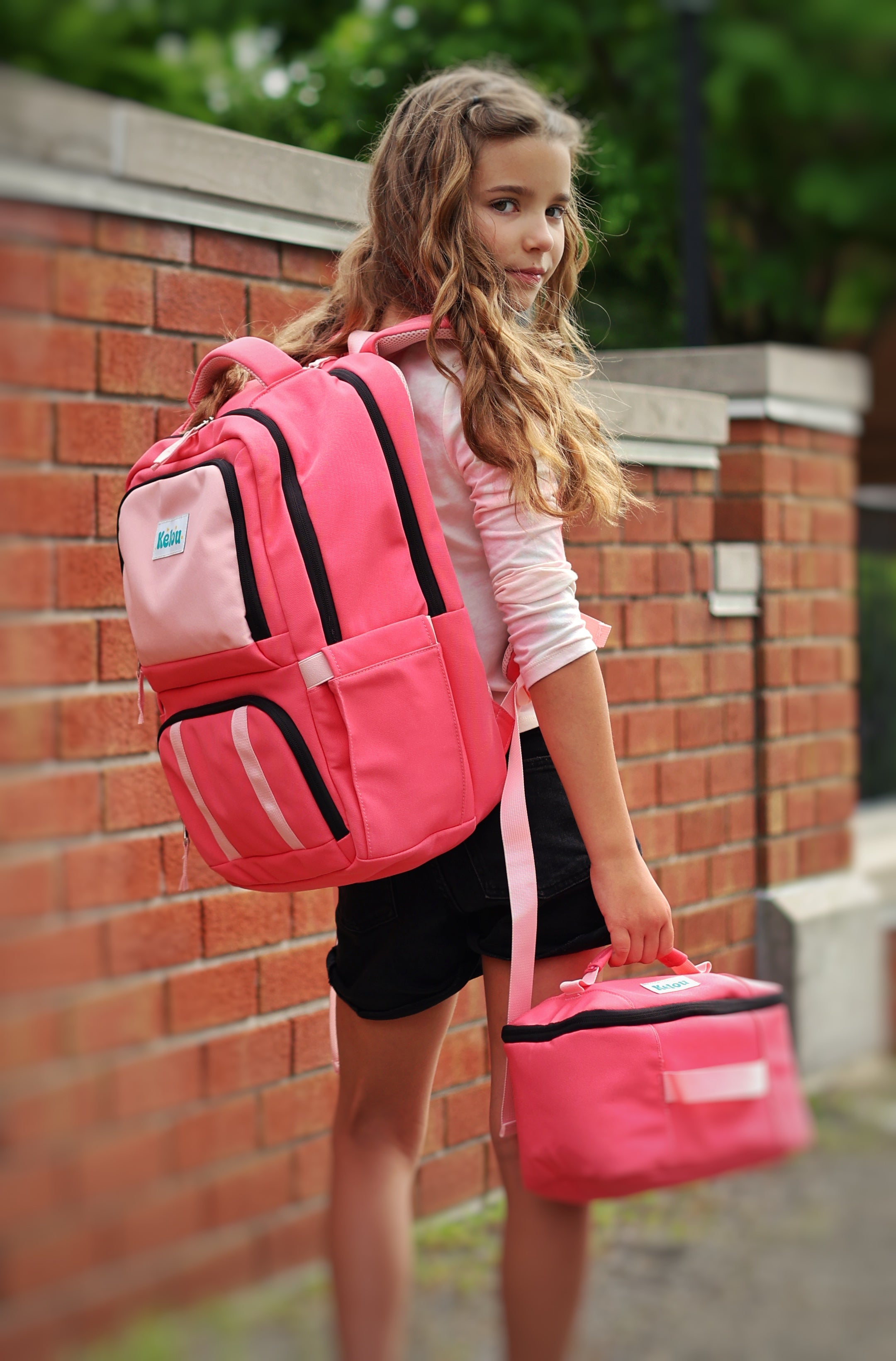 Girl with pink backpack and lunch bag standing against a brick wall