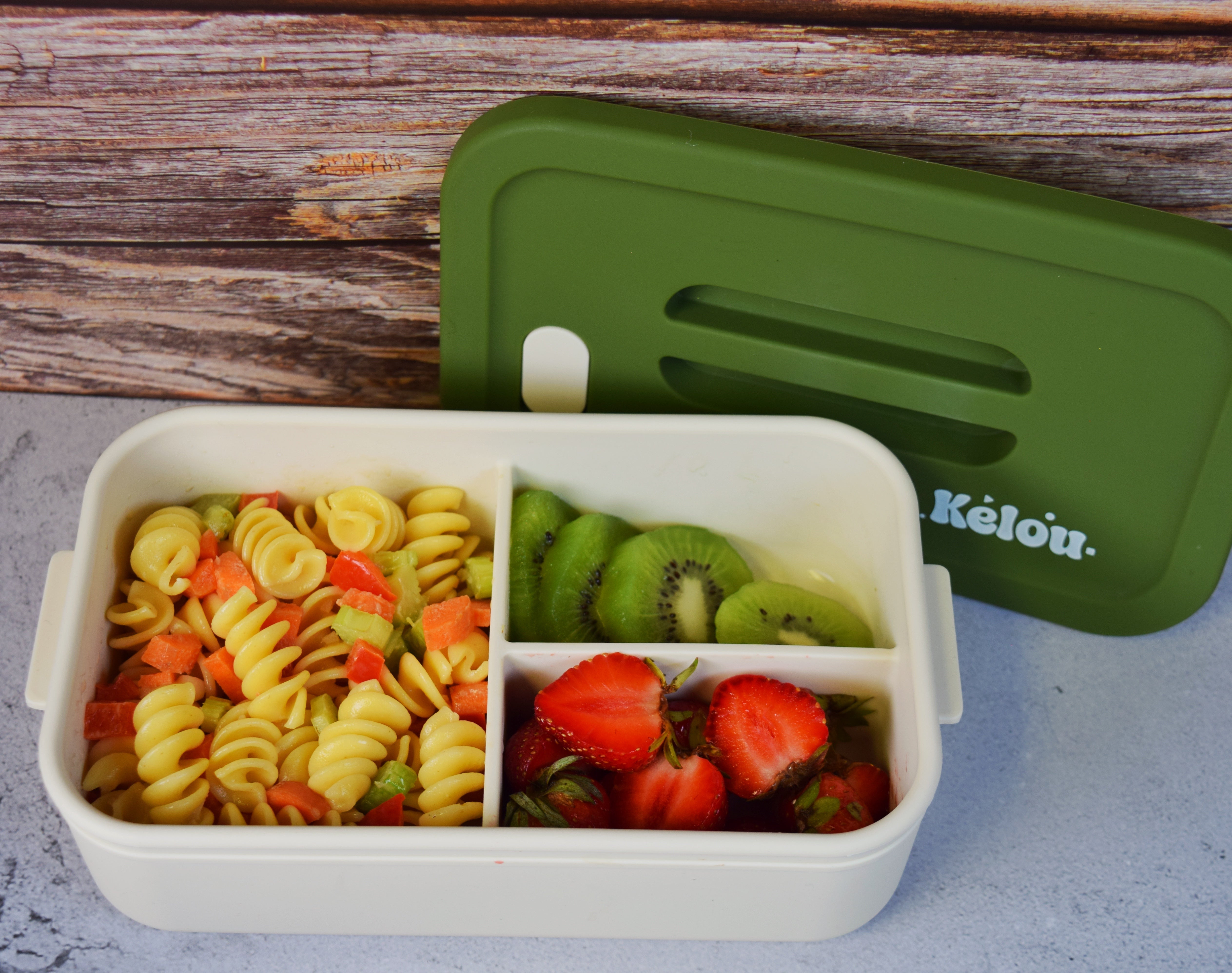 Bento box with pasta, kiwi, and strawberries next to a green lid on a wooden surface