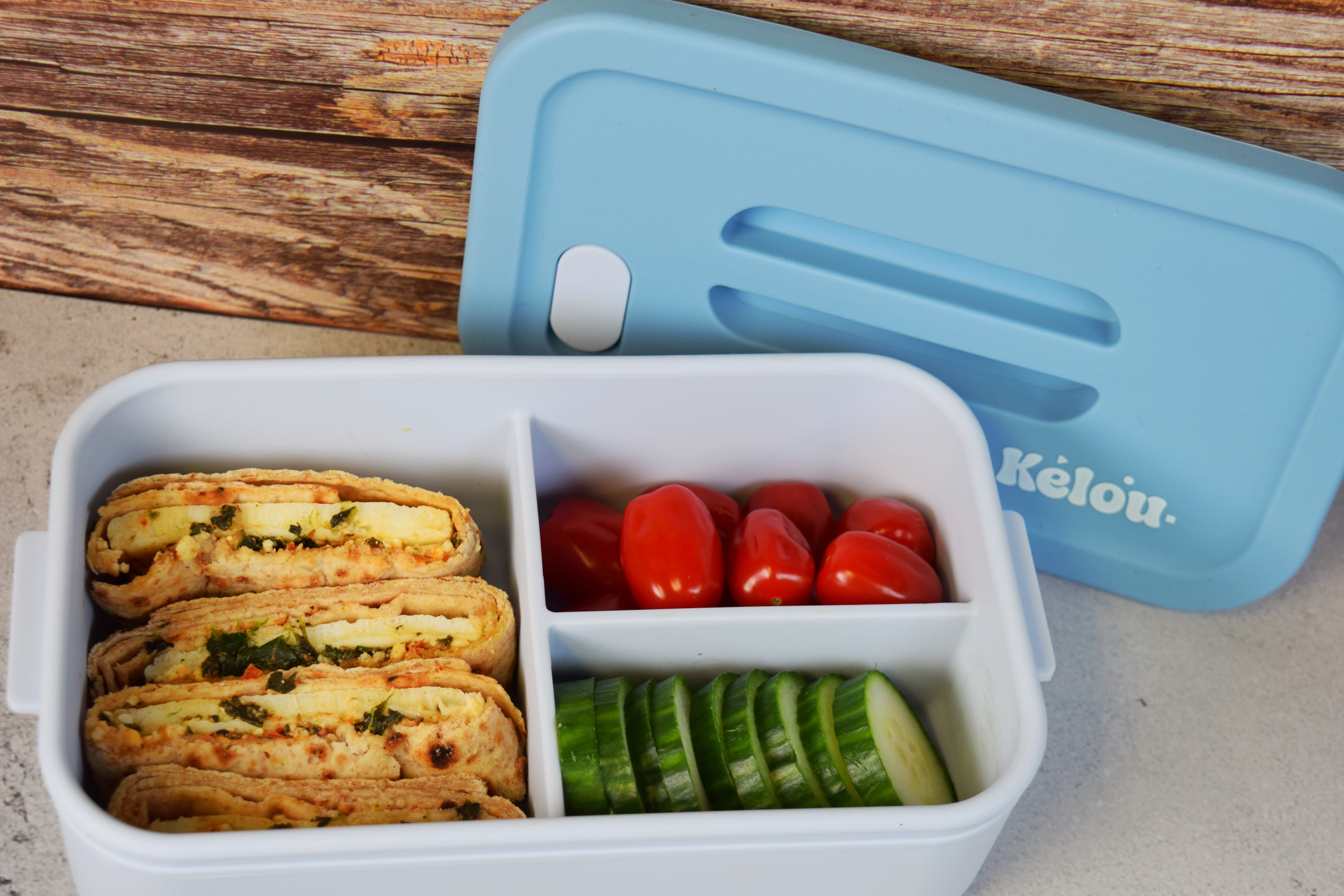 Bento box with food compartments on a wooden surface, featuring the brand 'Kelou'.