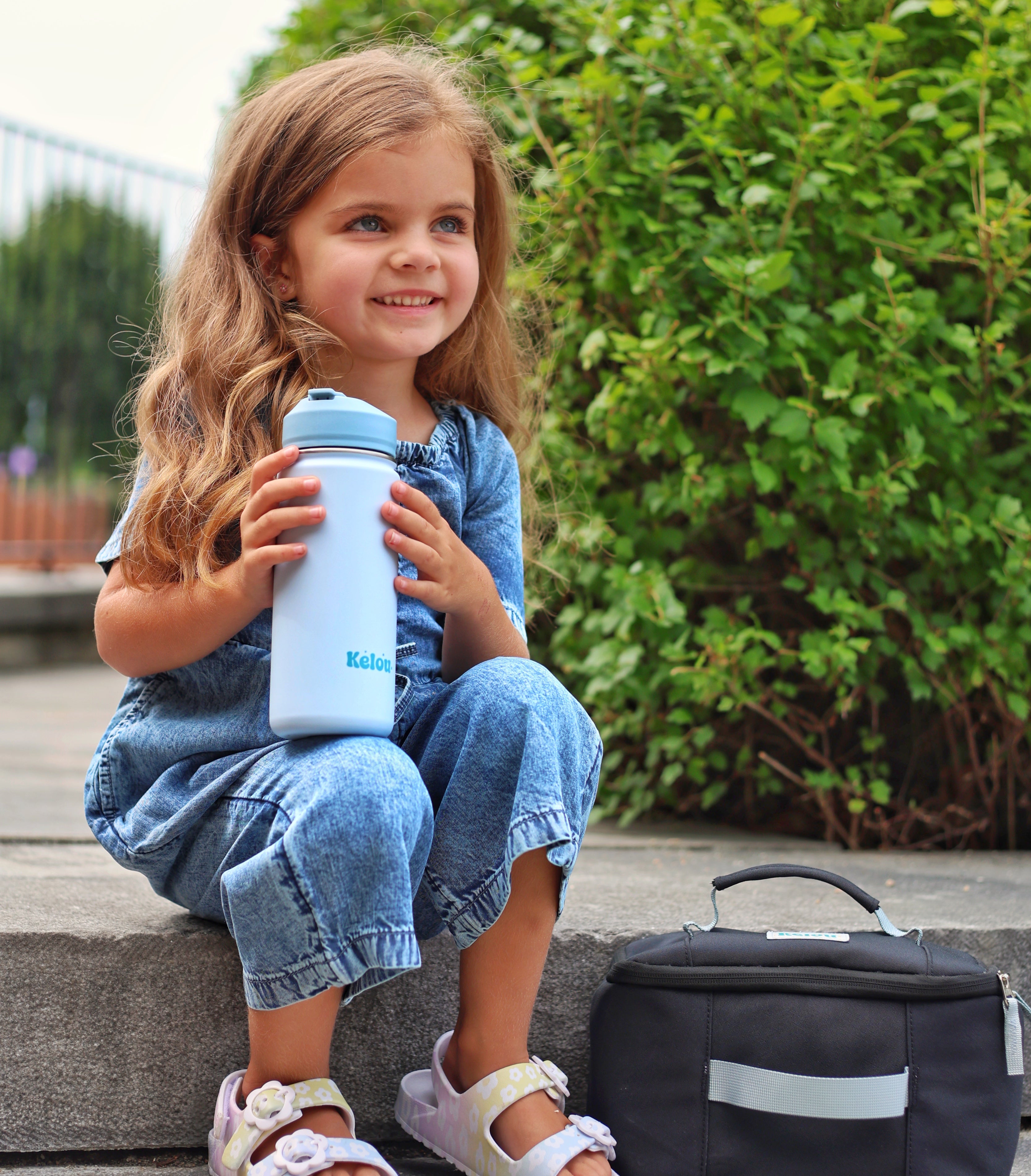 Child holding a blue water bottle with a black bag on a stone step.